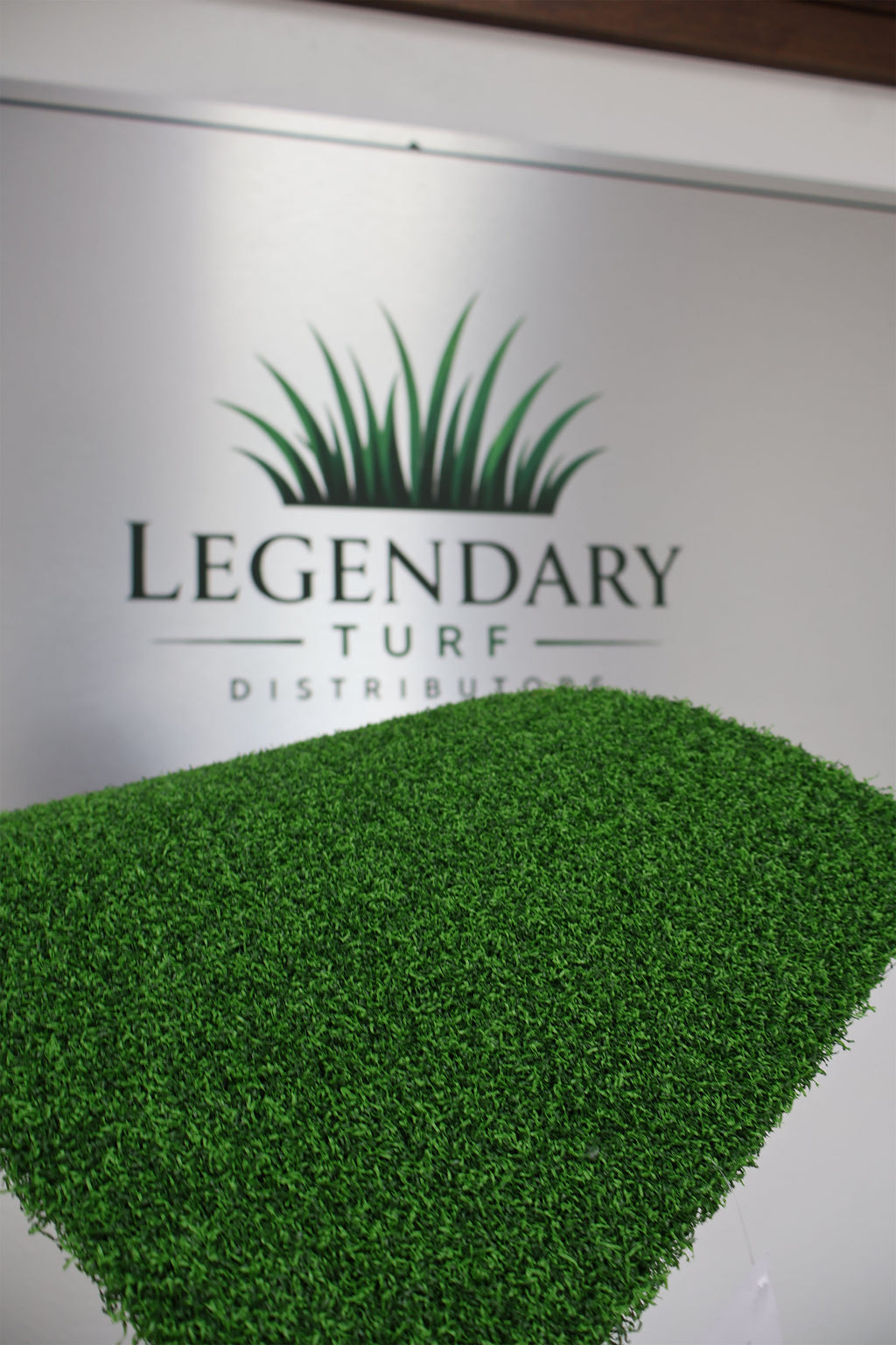 A close-up of bright green artificial turf or grass-like material in a diamond shape, placed in front of a white wall with the visible brand name Legendary Turf Distributors and a grass icon.
